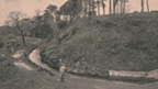 A small figure stands on a footpath by a narrow man-made water course at the foot of a hill with trees around.