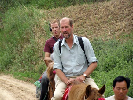 Presenter Martin Palmer and producer Mark O'Brien on horseback with unidentified guide, travelling along a dirt road through countryside