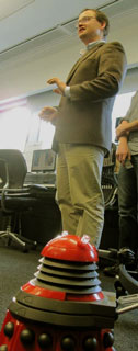 small red Dalek in front of man in brown jacket