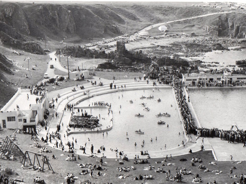 Black and white view, looking down from coastal cliffs to twin open air pools, with an Art Deco pavilion to the left of frame. One pool contains a mix of people paddling and rowing boats. The pool to the right is surrounded by a crowd of people, possibly watching a swimming competition. A large number of people are sunbathing on the surrounding grassy area.