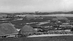 Black and white view of low, stone cottages with thatched roofs along the sides of a small loch. A larger, two-storey, pitched roofed building with slate or tile roof can be seen in the background.