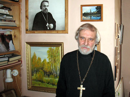 Father Alexander Borisov, a white-haired churchman, in his home with a photograph of Alexander Men visible on the wall behind him